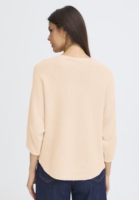 Luźny sweter w jasnym brzoskwiniowym kolorze, z rękawami do połowy przedramienia, prążkowaną fakturą i zaokrąglonym dołem. Połączony z ciemnoniebieskimi dżinsami.