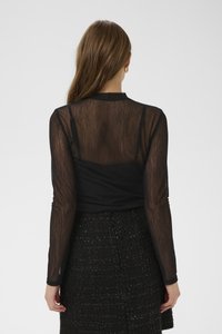 Moteris su ilgais rudais plaukais, dėvinti aukso žiedinius auskarus, permatomą juodą ilgomis rankovėmis viršūnę ant juodo kamisolės ir tekstūruotą juodą sijoną, žiūrinti į šoną.