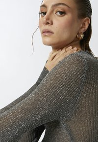 Metalik sivi, rebrast dugi rukav majica koja se odlikuje mekanom teksturom, izrezom u obliku suknje i slim fit krojem. Ruke modela odmaraju se na svom vratu.