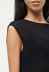 Femme portant un haut noir transparent sans manches, cadrée pour montrer l'épaule, le cou et les lèvres sur un fond blanc uni.