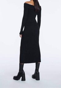 Abito nero a maglia, con spalle scoperte e maniche lunghe, dotato di spacco laterale e una silhouette aderente, abbinato a stivali neri con plateau alla caviglia.