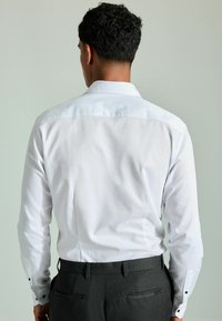 Bijela košulja dugih rukava s kragnom na dugme, s crnim dugmadi na manžetama, u kombinaciji s tamnosivim hlačama teksturiranog izgleda.