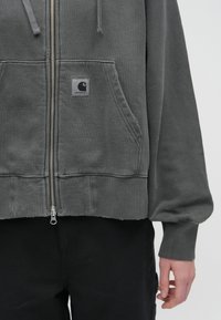 Sudadera con cremallera de algodón gris, con bolsillos delanteros, puños acanalados y un dobladillo recortado. Presenta un pequeño parche con el logo en el bolsillo izquierdo.