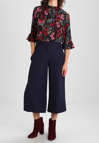 Top floreale a maniche lunghe con polsini arricciati in tonalità di blu navy e rosso, abbinato a culotte larghe blu navy e stivali alla caviglia bordeaux.