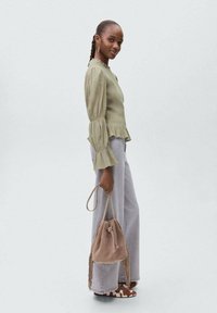 Svetlo zelena, strukturirana bluzica z nabranimi rokavi, v kombinciji z svetlo sivimi hlačami visoko pasu in bež nahrbtnikom s frfotajočimi robovi.