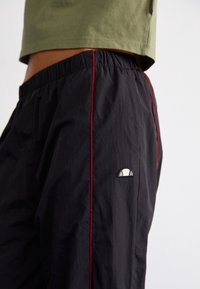 Pantalons de survêtement noirs avec une texture brillante, ornés de vidéos latérales rouges et d'un petit logo. Taille élastique pour un ajustement confortable.