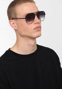 Lunettes de soleil noires avec verres dégradés, fine monture en métal et forme ronde, portées par une personne en t-shirt noir, sur un fond uni.