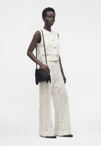 Femme portant un top blanc sans manches brodé et un pantalon large assorti, tenant un sac bandoulière noir à franges, debout devant un fond uni.