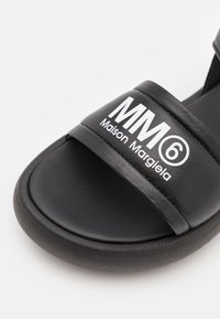 Svart slidesandal med en bred rem som har vit text "MM6 Maison Margiela." Materialet är glatt med en platt gummisula.