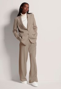 Beige op maat gemaakt pak met een blazer met één knoop en wijde broekspijpen, gecombineerd met een crèmekleurige coltrui en witte sneakers. Glad stofoppervlak.