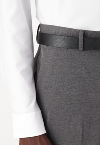 Pantalons gris en tissu texturé avec une ceinture en cuir noir. Poignet de chemise blanche visible, affichant une finition soignée.