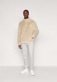 Beige, plyšový fleece pulover s polovičným zipsom a logom na hrudi, skombinovaný s svetlo šedými teplákmi a bielymi teniskami. Ploche pletené manžety na leme.