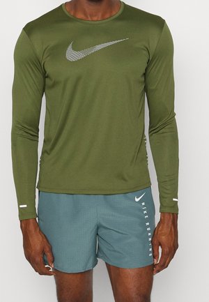 Mann trägt ein olivgrünes langärmliges Sportshirt mit weißem Nike-Logo und blaue perforierte Shorts mit Nike Running-Logo auf dem rechten Bein.