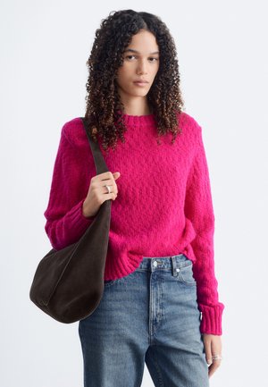 Jeune personne aux cheveux bouclés portant un pull rose vif et un jean bleu, portant un sac à bandoulière marron foncé.