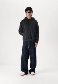 Haut à capuche noir avec une poche avant, associé à un jean en denim foncé ample. Le modèle porte des chaussures crème avec des semelles marron. Le mannequin porte des lunettes de soleil noires.