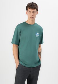 Grøn kortærmet t-shirt lavet af bomuld, prydet med et farverigt bjergdesign på venstre bryst. Sammen med mørke ternede bukser.