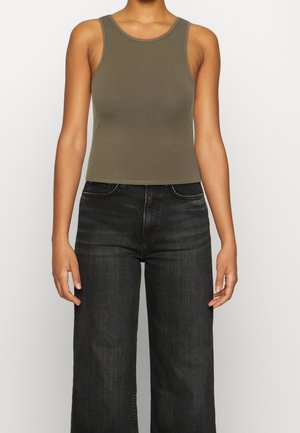 Personne portant un débardeur court olive sans manches et un jean noir taille haute à jambes larges, debout devant un fond uni.