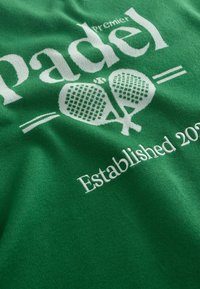 Tissu vert avec texte blanc « premier padel » présentant deux raquettes de padel croisées et « Established 2023 » en dessous.