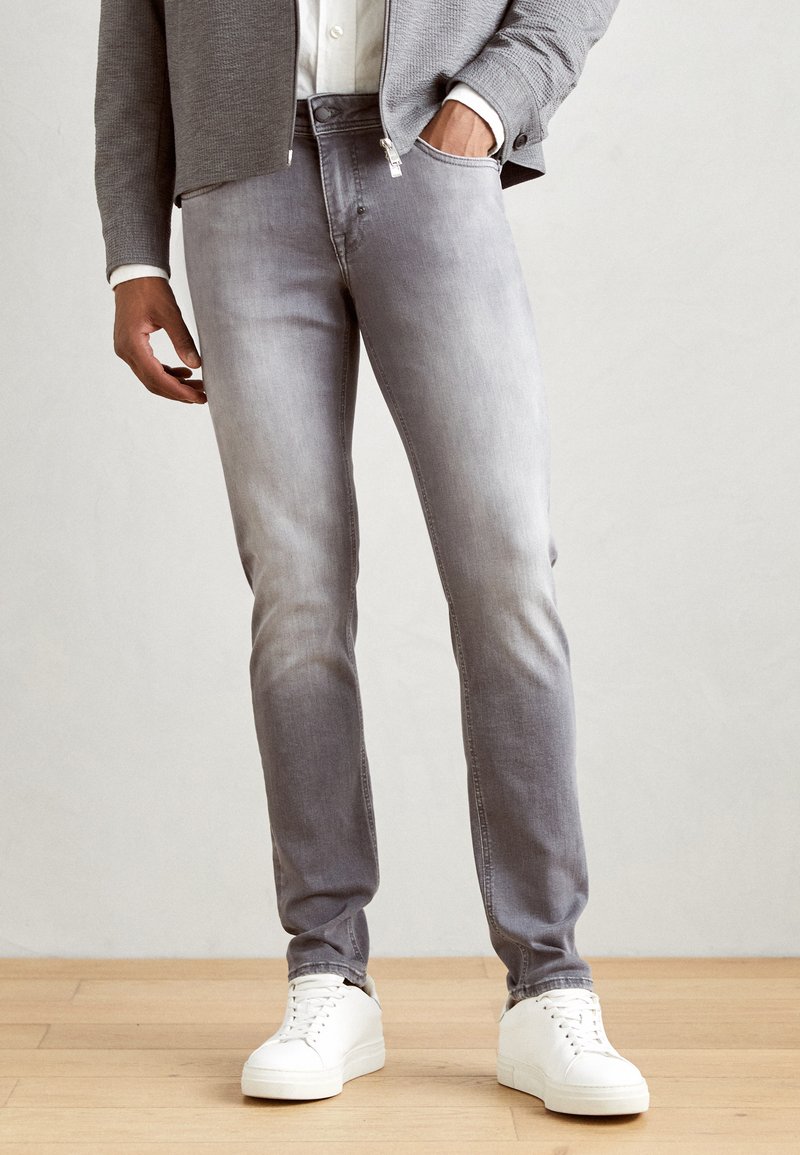 Jeans de denim gris con un ligero desgaste, corte slim y diseño de cinco bolsillos, combinados con zapatillas blancas sobre un fondo de suelo de madera.