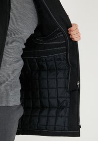 Černá prošívaná podšívka vnitřní bundy se šedou žebrovanou vnější vrstvou. Má viditelné švy a boční kapsy. Zapínání na zip.