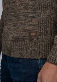Hnědý pletený svetr s texturovanými vzory, pružnými manžetami a spodním lemem, s malou embosovanou koženou nášivkou na přední dolní straně.