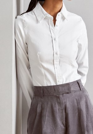 Osoba w białej koszuli na guziki wsuniętej w szare, plisowane spodnie z wysokim stanem, stojąca na tle jasnej ściany.