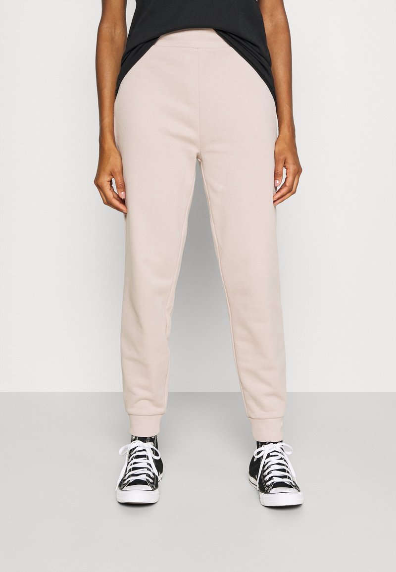 Helle beige Sweatpants mit elastischem Bund, seitlichen Taschen und gerippten Bündchen, kombiniert mit schwarz-weißen Sneakern. Glatte Textur.