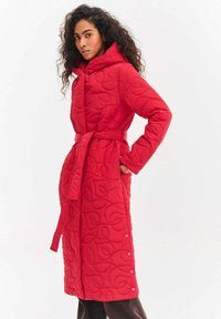Czerwony pikowany długi płaszcz z kapturem, paskiem w talii i teksturowanym wzorem. Posiada duże guziki przy dolnym brzegu oraz gładkie wykończenie.