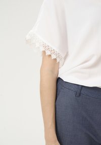 Bela bluza z čipkastim robom na rokavih v kombinaciji s temno modrimi hlačami. Bluza je gladka, medtem ko čipka vsebuje cvetlične vzore.