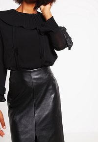 Blusa negra plisada con un alto cuello rizado y mangas acampanadas con volantes, combinada con una falda de cuero negra ajustada con una abertura.