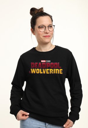 Kvinde med briller iført en sort sweatshirt med Marvel Studios Deadpool og Wolverine logo, står mod en ensfarvet lys baggrund.