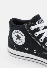 Svarta hög-top sneakers av canvas med vita snören, en gummisula och en rund logotyp-patch som har en stjärna och texten "Converse All Star."