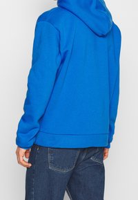 Sudadera azul con capucha y cordón, confeccionada en tela suave. Presenta un corte holgado y puños acanalados, combinada con vaqueros de mezclilla oscura.