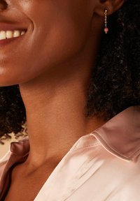 Des boucles d'oreilles en or avec un design de chaîne présentent une perle rose ronde suspendue à l'extrémité. Le modèle a les cheveux bouclés et porte une chemise rose clair.