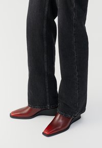 Persona che indossa jeans neri a gamba larga e stivaletti in pelle rossa con punta quadrata e tacco basso su uno sfondo bianco.