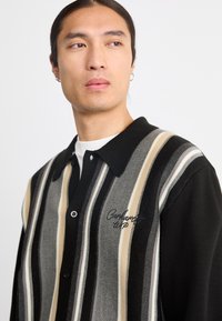 Maglione a righe in nero, grigio e crema, con colletto nero, chiusura con bottoni e logo ricamato sul petto.