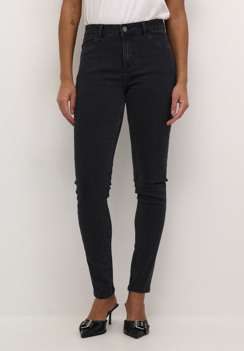 Jean skinny noir taille haute avec une texture lisse, présentant une fermeture à bouton à l'avant et cinq poches, associés à des mules à talons noires.