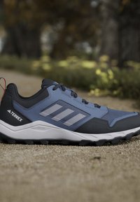 Chaussure de trail running Adidas Terrex bleue et noire sur du gravier avec une semelle robuste et un arrière-plan en plein air avec des arbres et du feuillage.
