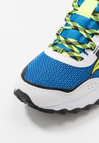 Blaue Sportschuh mit einem strukturierten Mesh-Obermaterial, weißen und schwarzen Akzenten sowie einem gemusterten neon-gelben und schwarzen Schnürsystem.