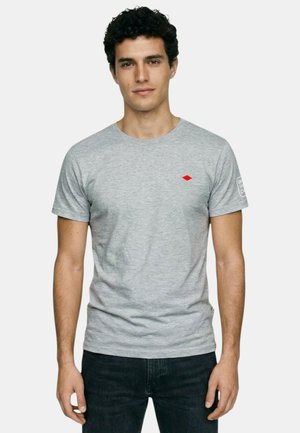 Homme aux cheveux courts, bouclés et foncés, portant un T-shirt à manches courtes gris clair avec un logo en forme de losange rouge et un pantalon noir, debout devant un fond blanc.