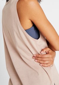 Top senza maniche beige chiaro con tessuto morbido e testurizzato e una vestibilità ampia, che presenta un design della schiena arrotondato e un'imbottitura blu visibile.