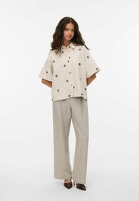 Mujer con una camisa beige de botones con patrones negros, pantalones beige sueltos y tacones oscuros de punta, de pie con las manos detrás de la espalda.