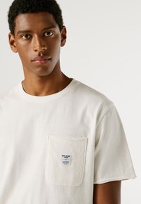 Valge puuvillane T-särk lühikeste käistega, ümmarguse kaelusega ja vasakul rinnal taskuga, millel on logotähis. Pehme tekstuur, argine disain.