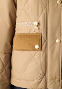 Béžová prošívaná bunda s kapsou na zip se zlatými doplňky a akcentem z manšestru na přední části. Kulaté logo na patentkách.