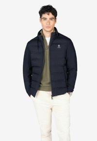 Chaqueta acolchada azul marino con cierre frontal, que presenta secciones cosidas y un logo en el pecho, llevada sobre un suéter verde oliva y pantalones claros.