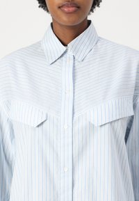 Camicia button-up a righe blu chiaro con colletto, due tasche sul petto e vestibilità ampia. Il materiale sembra morbido con una superficie liscia.