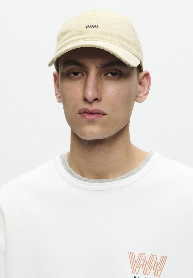 Beige corduroy cap with embroidered "W.W." logo. Model wears a white sweater with gray accents and an orange graphic on the chest.