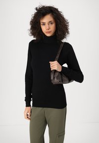 Crna turtleneck pulover, uski dizajn, rebraste manžete. Maslinasto zelene hlače s detaljima na zip-u. Drži malu smeđu kožnu torbicu.
