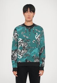 Felpa turchese con accenti neri, caratterizzata da intricati disegni in bianco e nero di animali, oggetti ed elementi decorativi. Maniche lunghe.