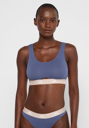 Woman wearing blue sports bra and matching underwear with beige Adidas logo bands, standing against a plain light background.